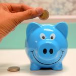 pexels-photo-1602726-1602726 Hand inserting a coin into a blue piggy bank for savings and money management.